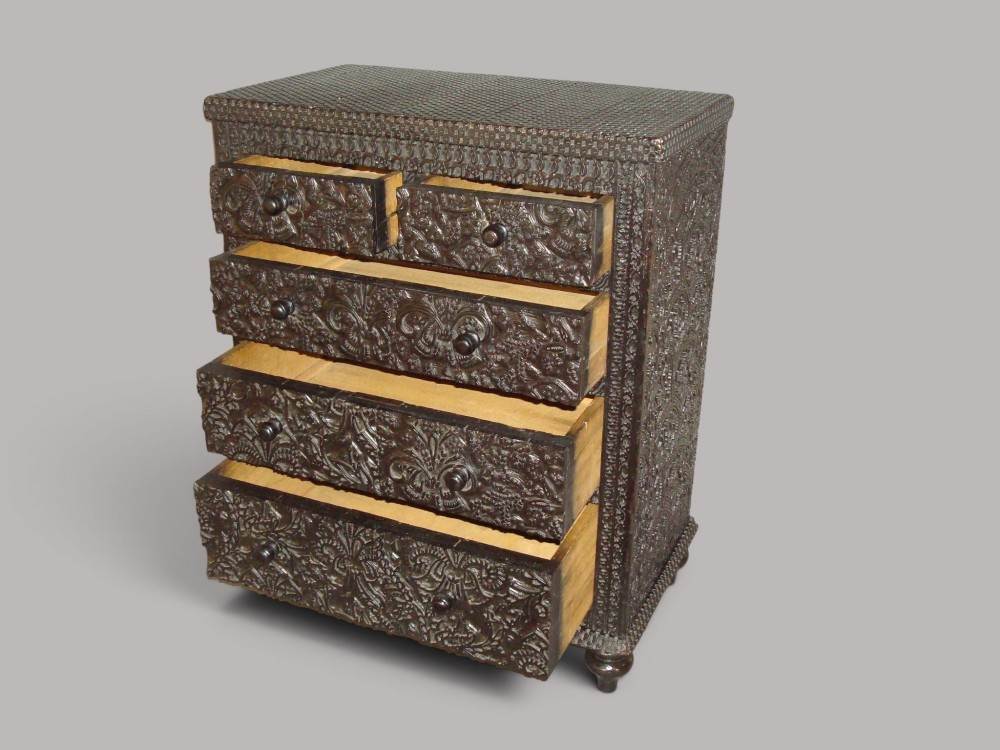 A 19th-century small ebonised chest of drawers featuring relief foliage carving across all surfaces, turned wooden knobs, and short bun feet. View X
