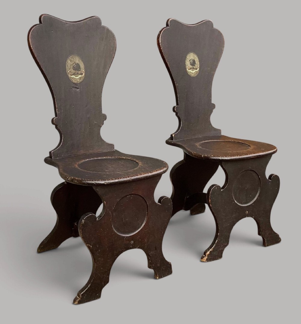 A pair of dark brown 18th-century Irish oak sgabello hall chairs with shaped backs featuring painted armorial crests.