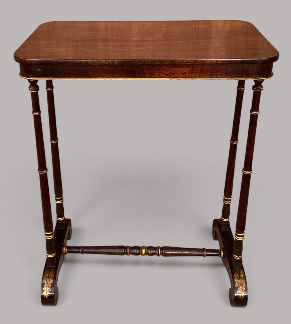A Regency period dark rosewood occasional table featuring a rectangular top with rounded corners, supported by twin turned spindle legs, a central stretcher, and parcel-gilt foliage decoration on the feet.