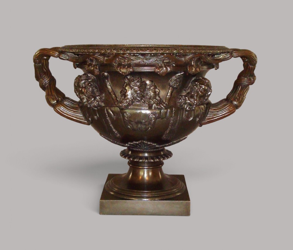 A 19th-century patinated bronze Warwick vase with relief Bacchanalian masks, intertwined vine handles, and a square base.
