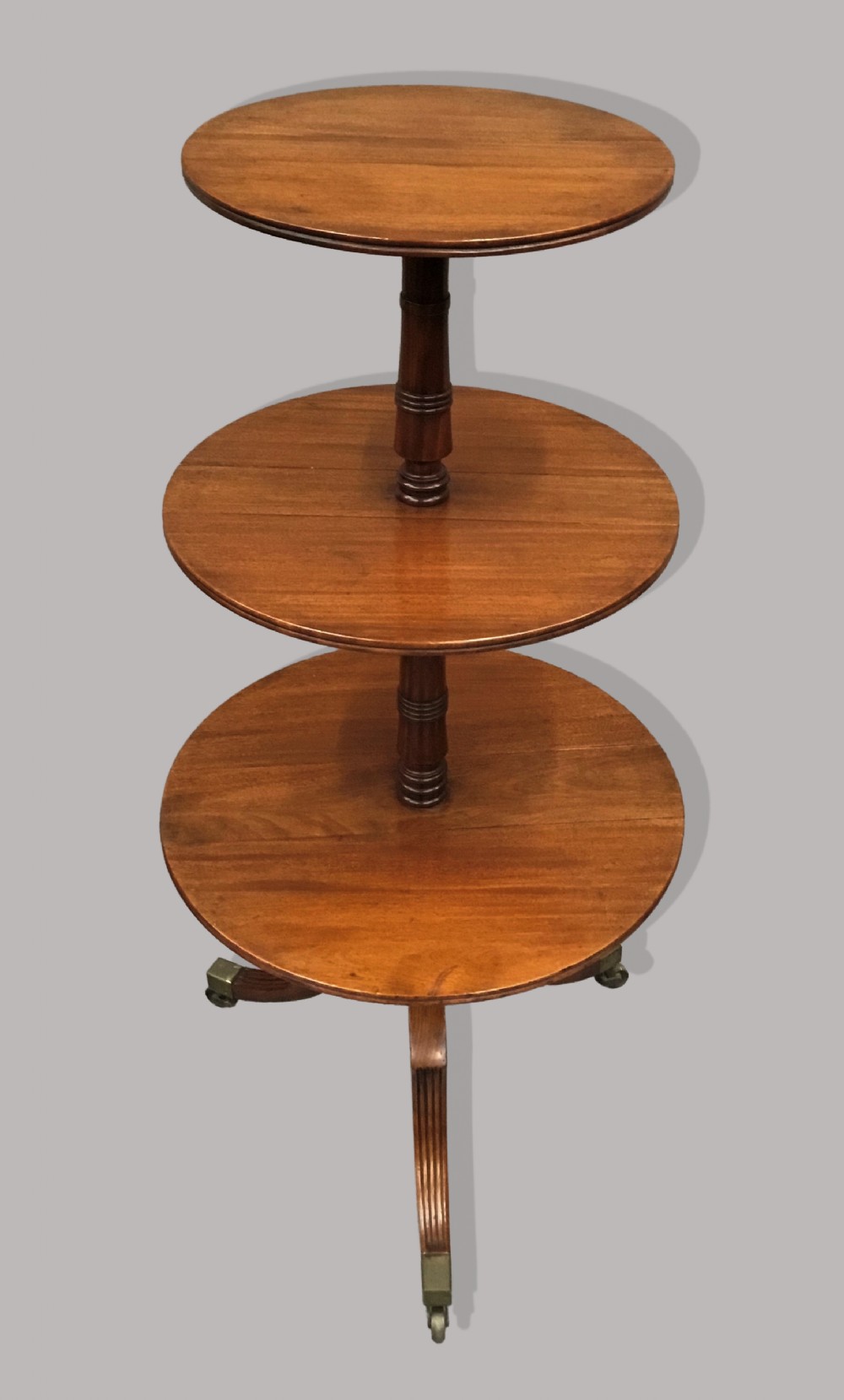 A George III mahogany three-tier dumb waiter featuring circular graduated folding surfaces, turned central support columns, and tripod legs with brass castors.