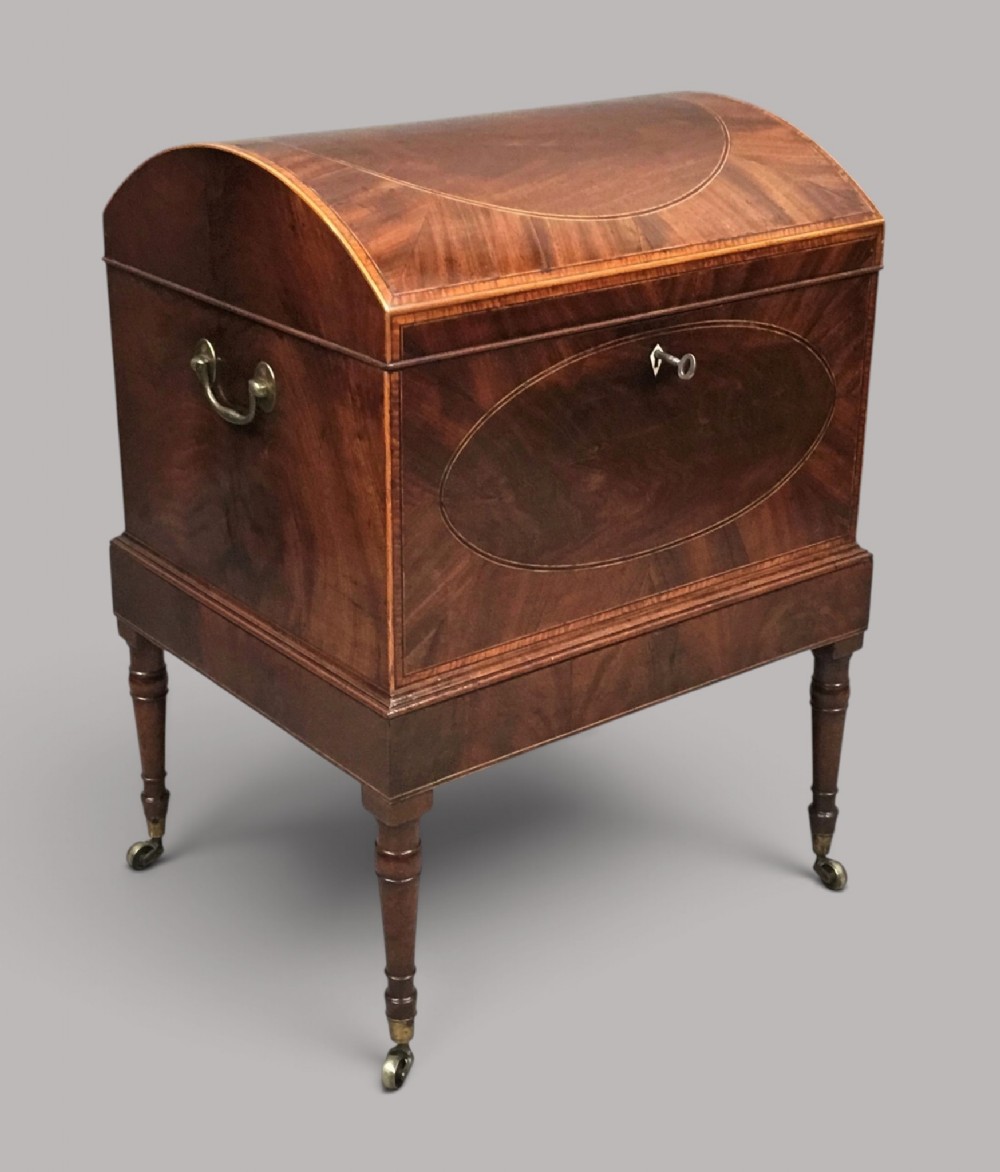A George III Sheraton style flame mahogany cellarette featuring a domed lid, oval stringing inlay, brass side handles, and turned legs on castors.
