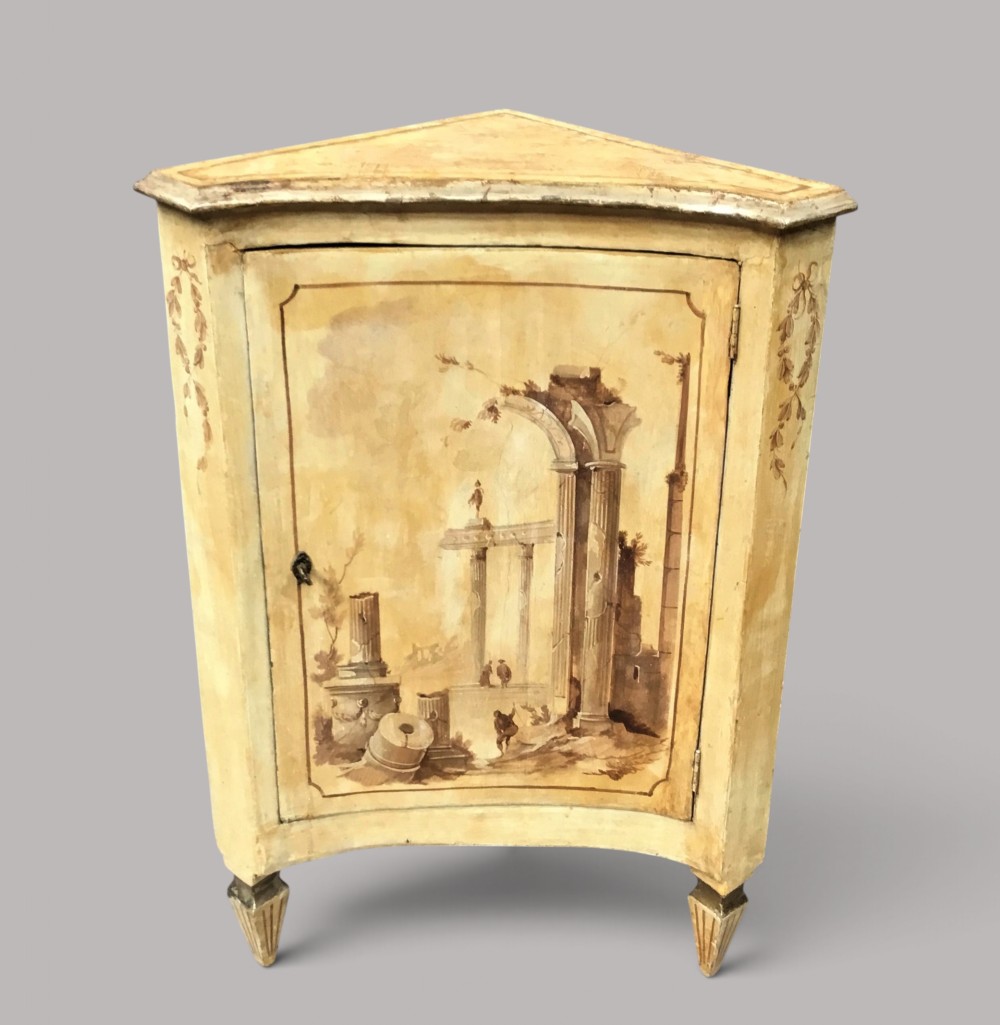 An 18th-century cream-painted Italian standing corner cabinet featuring a curved door decorated with a painted architectural scene and tapered legs.