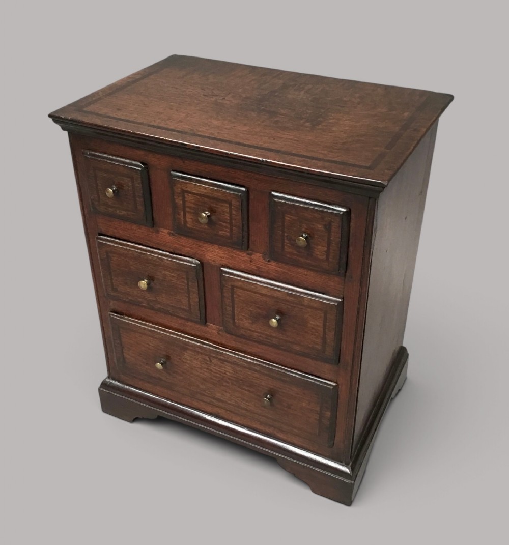 An 18th-century dark oak miniature chest of drawers featuring crossbanded inlay, six drawers with brass knobs, and bracket feet.