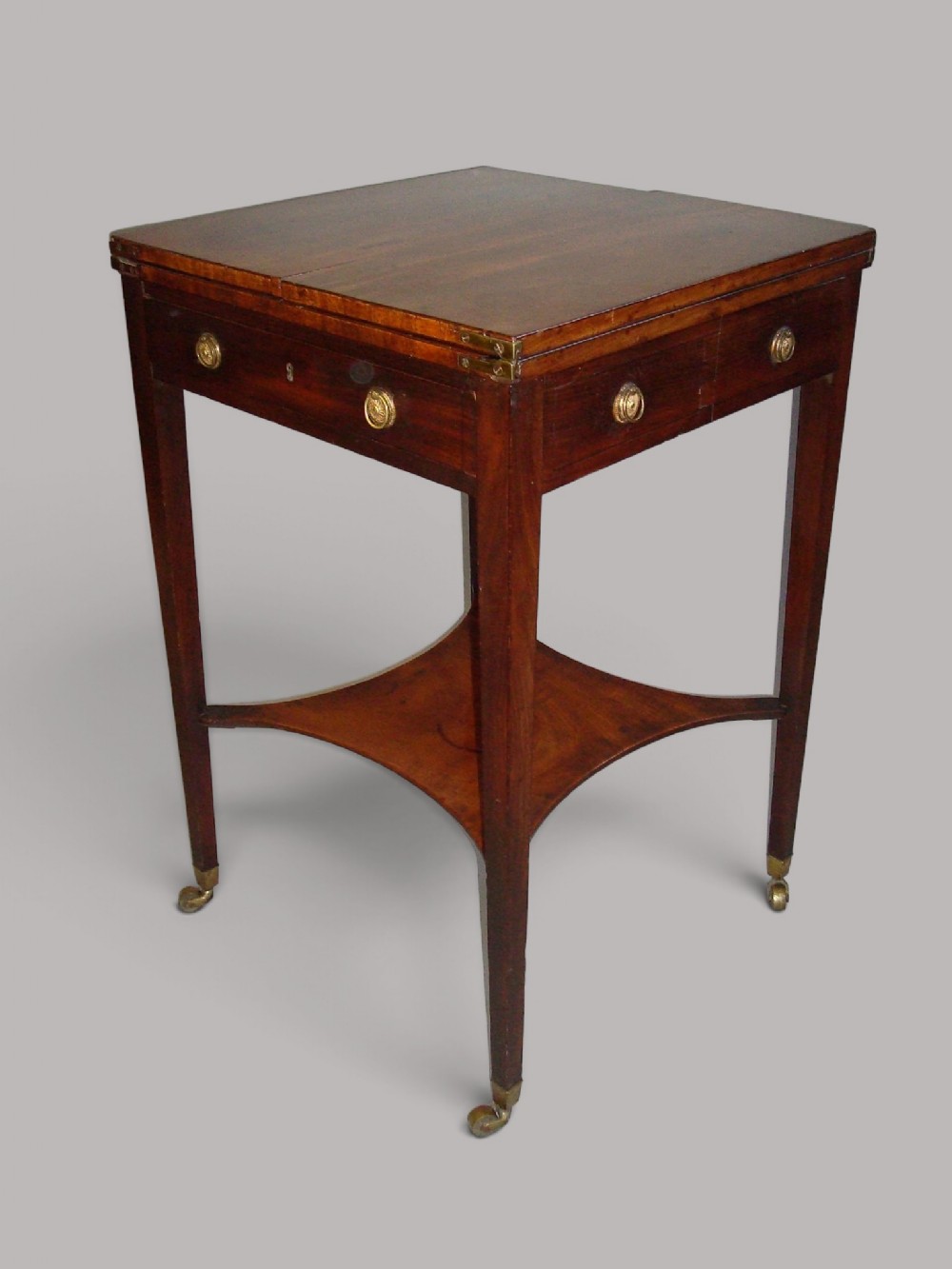 A George III mahogany patience table featuring a hinged folding top, brass hardware, a concave lower shelf, and tapered legs with brass castors.