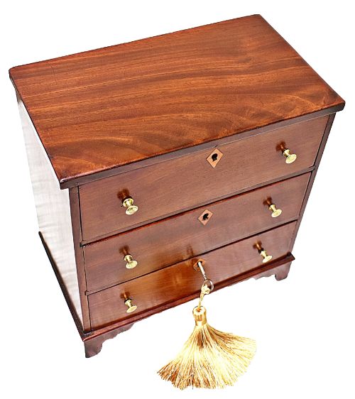 fig 2. A Regency period miniature mahogany chest featuring three stacked drawers, small brass knobs, diamond escutcheons, shaped bracket feet, and a key with a tassel.
