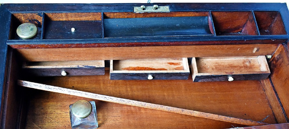 An open antique mahogany writing slope featuring a red gold-tooled leather writing surface, brass corner mounts, and dedicated compartments for inkwells. View X