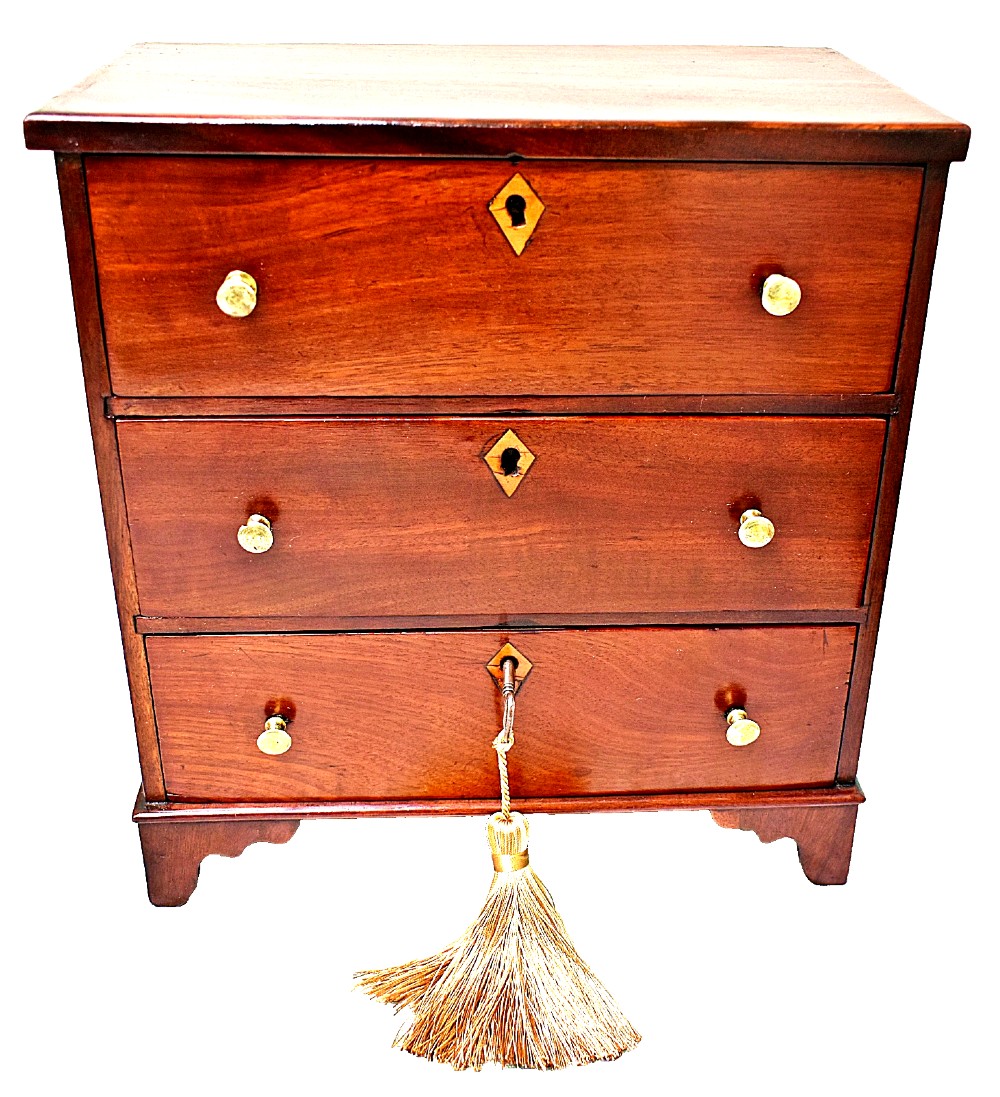 A Regency period miniature mahogany chest featuring three stacked drawers, small brass knobs, diamond escutcheons, shaped bracket feet, and a key with a tassel.