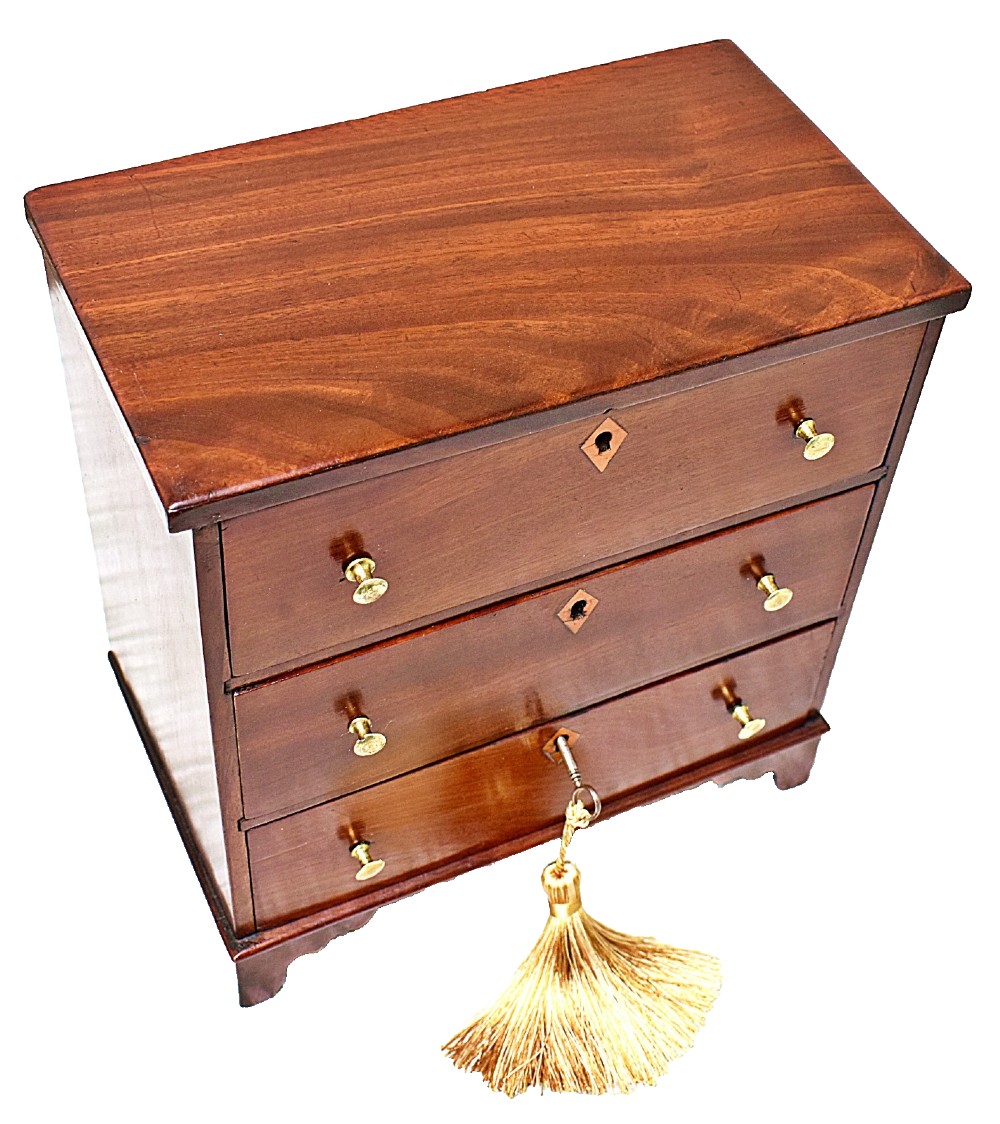 A Regency period miniature mahogany chest featuring three stacked drawers, small brass knobs, diamond escutcheons, shaped bracket feet, and a key with a tassel. View III