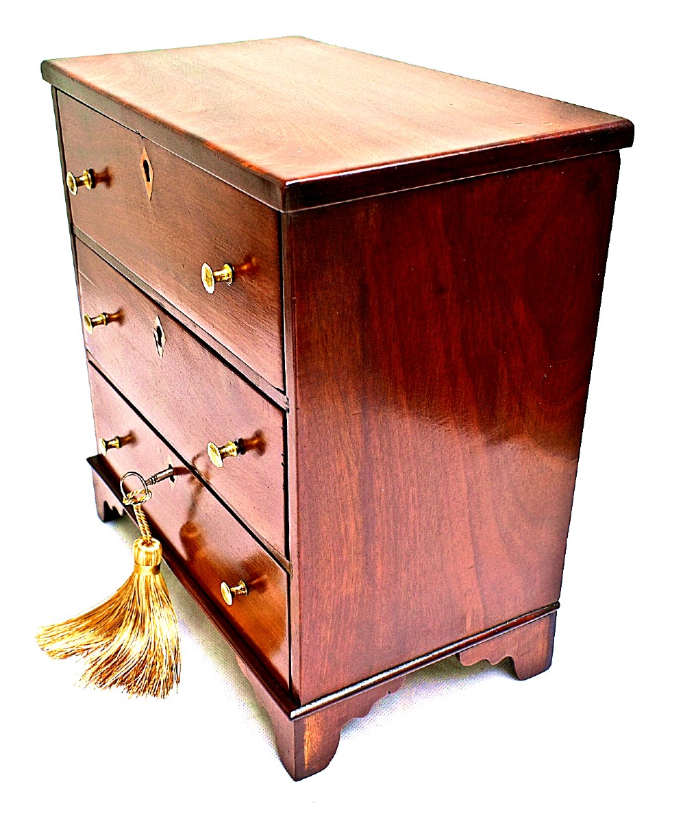 A Regency period miniature mahogany chest featuring three stacked drawers, small brass knobs, diamond escutcheons, shaped bracket feet, and a key with a tassel. View IV