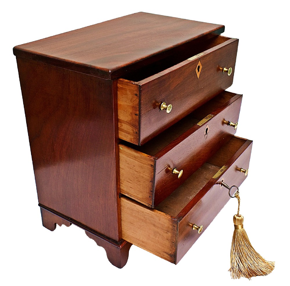 A Regency period miniature mahogany chest featuring three stacked drawers, small brass knobs, diamond escutcheons, shaped bracket feet, and a key with a tassel. View VI