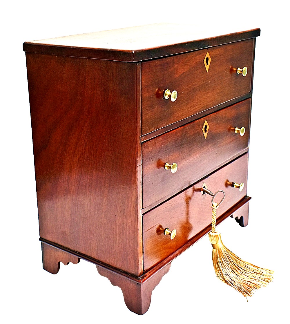 A Regency period miniature mahogany chest featuring three stacked drawers, small brass knobs, diamond escutcheons, shaped bracket feet, and a key with a tassel. View IX