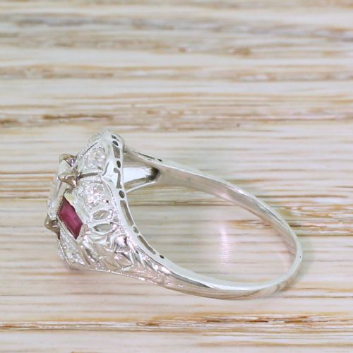 fig 2. A retro silver-tone ring featuring a central emerald-cut diamond flanked by two rectangular rubies surrounded by pierced filigree work.