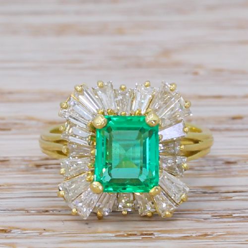 fig 1. A yellow gold ballerina ring featuring a central rectangular emerald cut green emerald surrounded by a flared halo of baguette diamonds.