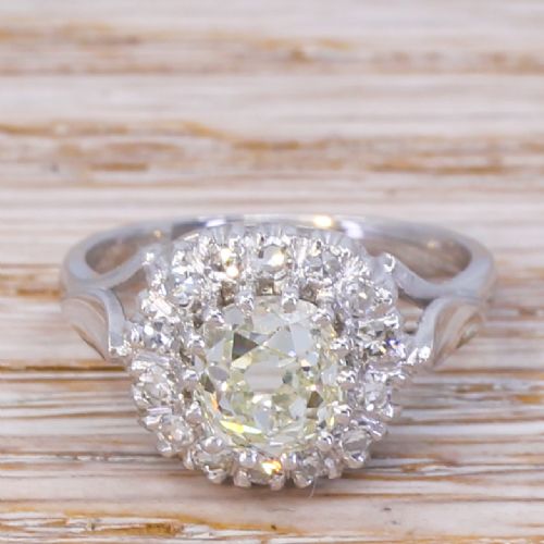 fig 1. An Art Deco style white metal ring featuring a central round diamond surrounded by a halo of smaller diamonds on a carved shoulder band.