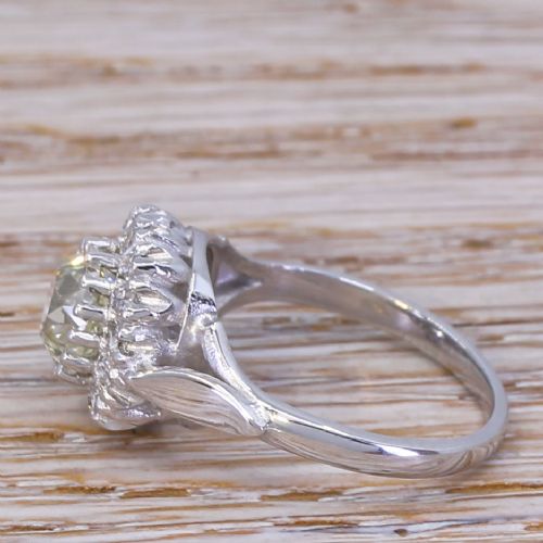 fig 2. An Art Deco style white metal ring featuring a central round diamond surrounded by a halo of smaller diamonds on a carved shoulder band.