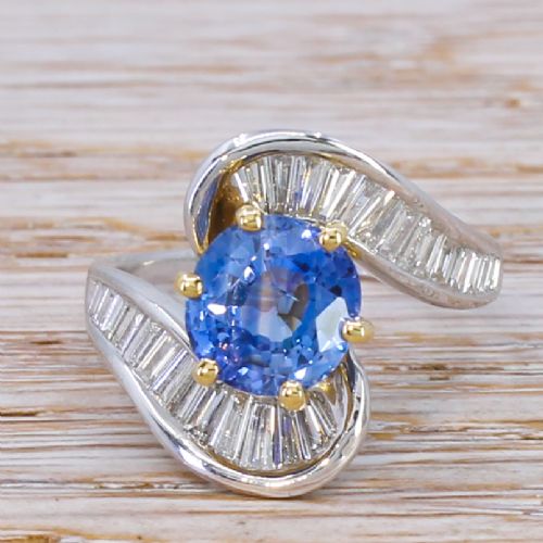 fig 1. Mid Century crossover ring featuring a round blue sapphire mounted in yellow gold claws flanked by channel-set baguette diamonds in a sweeping wave band.