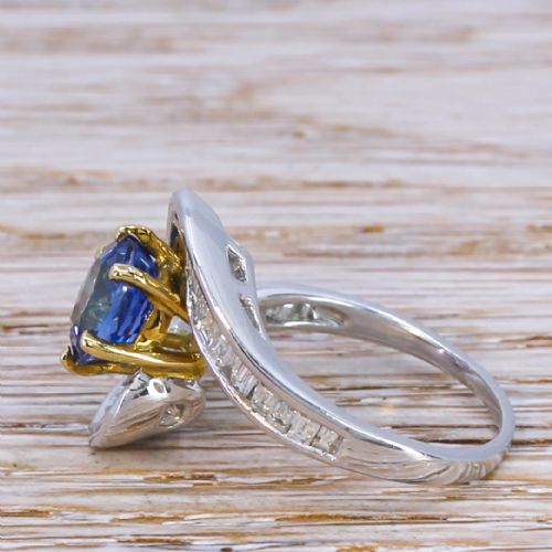 fig 2. Mid Century crossover ring featuring a round blue sapphire mounted in yellow gold claws flanked by channel-set baguette diamonds in a sweeping wave band.
