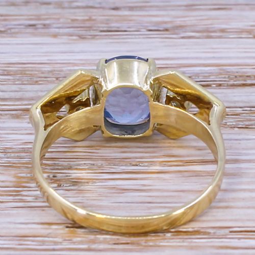 fig 3. A yellow gold ring featuring a central oval-cut light blue sapphire flanked by two round diamonds within geometric gold shoulders.
