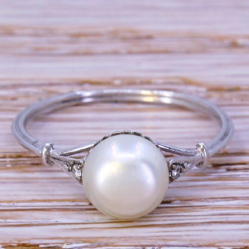 fig 1. A mid-century ring featuring a smooth, light cream natural pearl solitaire mounted on a slender silver-coloured band with subtle diamond accents on the shoulders.