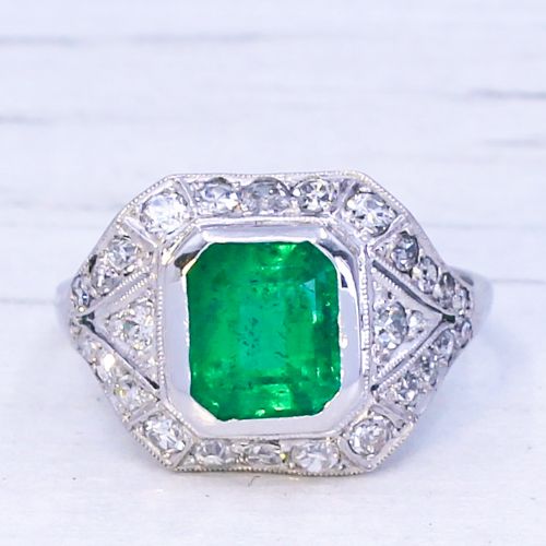 fig 2. An Art Deco platinum ring featuring a central rectangular green emerald surrounded by a geometric halo of white diamonds with milgrain detailing.
