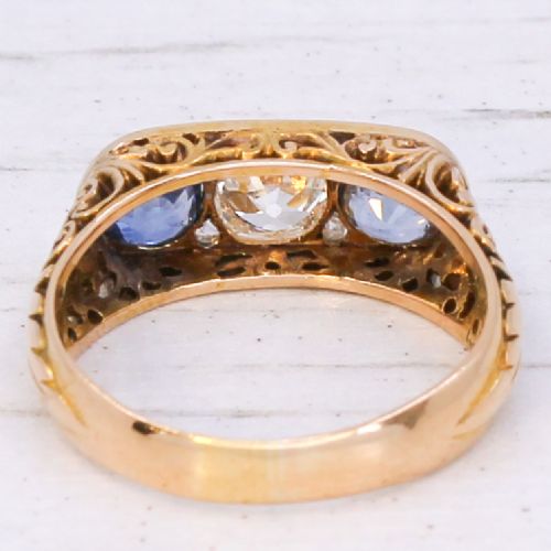 fig 4. An Art Nouveau yellow gold ring set with a central round old cut diamond flanked by two blue sapphires within an openwork scrollwork gallery.