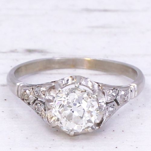 fig 2. An Art Deco style white metal engagement ring featuring a central round old cut diamond flanked by smaller accent diamonds on sculpted shoulders.