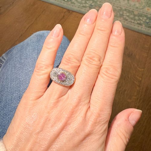 fig 1. A vintage gold ring featuring a central bezel-set pink sapphire flanked by two round diamonds within a pave-set diamond oval plaque.