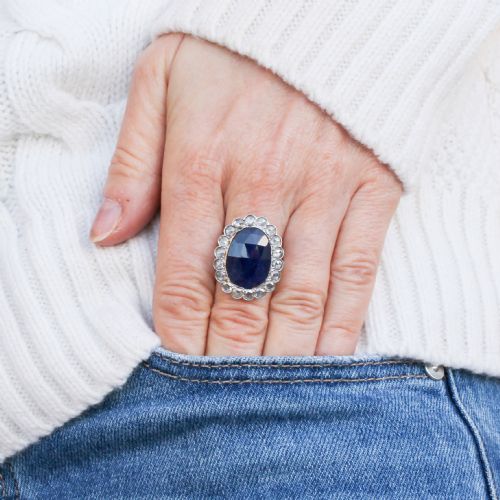 fig 1. An oval blue sapphire gemstone set in a silver-toned metal ring, surrounded by a halo of circular white diamonds.