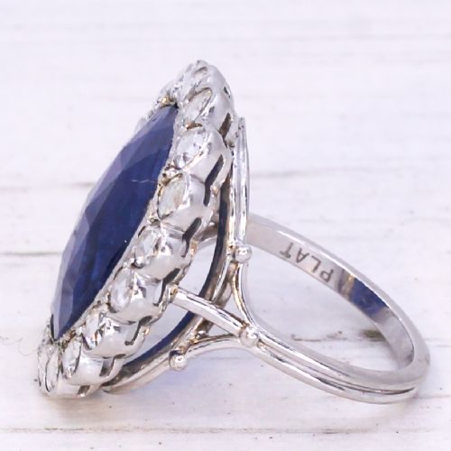 fig 3. An oval blue sapphire gemstone set in a silver-toned metal ring, surrounded by a halo of circular white diamonds.