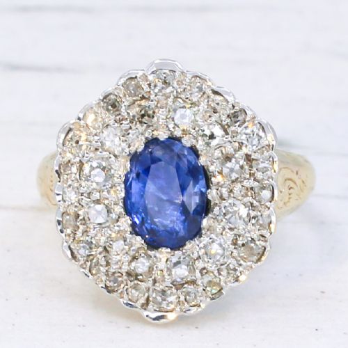 fig 2. An oval blue sapphire surrounded by a double halo of diamonds set in a scalloped white metal mount on an engraved yellow gold band.
