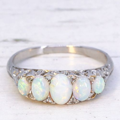 fig 2. A white metal ring featuring five graduated oval cabochon opals set in a row across a decorative scrollwork setting.