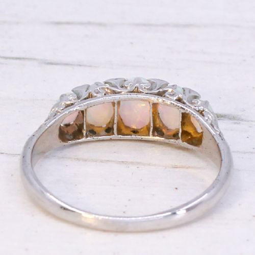 A white metal ring featuring five graduated oval cabochon opals set in a row across a decorative scrollwork setting. View V