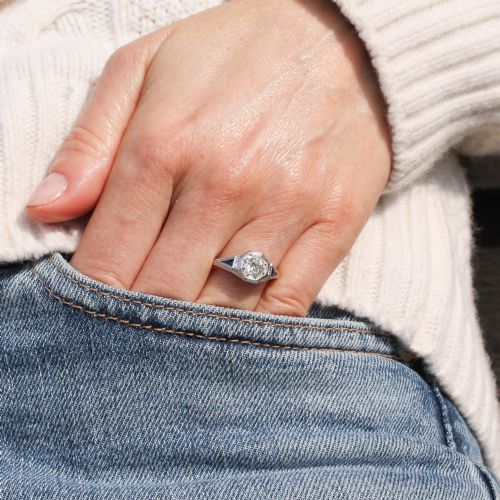 fig 1. A white metal ring featuring a central old-cut diamond set within an octagonal bezel, flanked by triangular blue sapphires and fine engraved detailing along the band.
