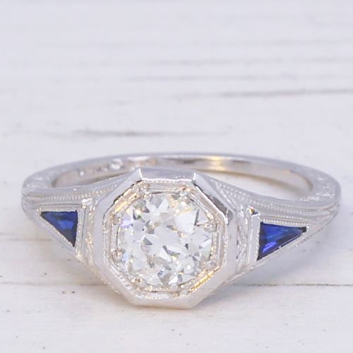 fig 2. A white metal ring featuring a central old-cut diamond set within an octagonal bezel, flanked by triangular blue sapphires and fine engraved detailing along the band.