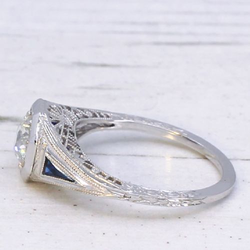 fig 3. A white metal ring featuring a central old-cut diamond set within an octagonal bezel, flanked by triangular blue sapphires and fine engraved detailing along the band.