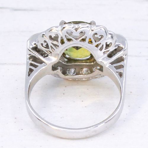 A vintage white precious metal ring featuring a central oval green sapphire encircled by a halo of round diamonds and diamond-set shoulders. View V