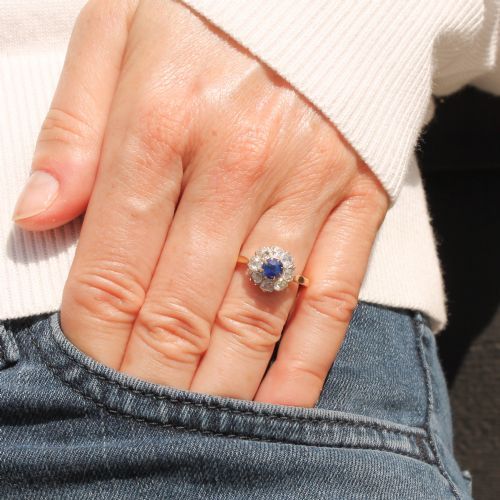 fig 1. A vintage cluster ring featuring a central round blue sapphire encircled by nine brilliant-cut diamonds set on a yellow gold band.
