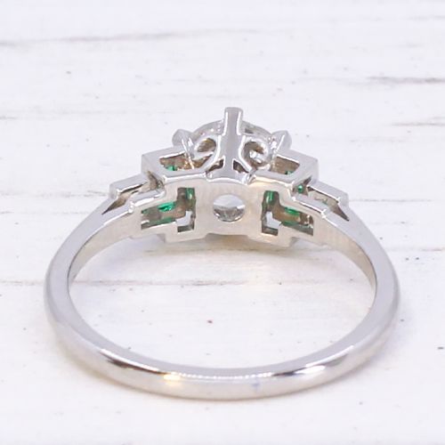 A platinum ring set with a central round old European cut diamond flanked by channel-set green emerald shoulders. View V