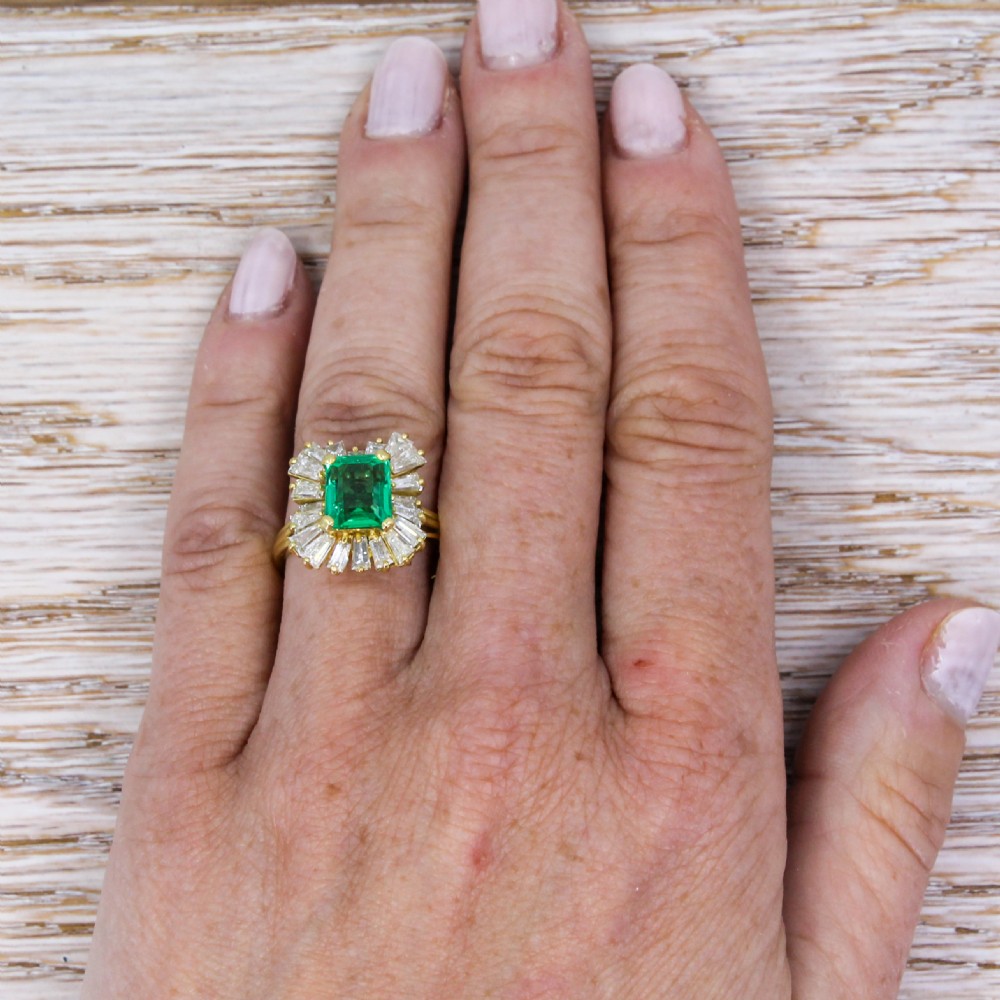 A yellow gold ballerina ring featuring a central rectangular emerald cut green emerald surrounded by a flared halo of baguette diamonds. View VI