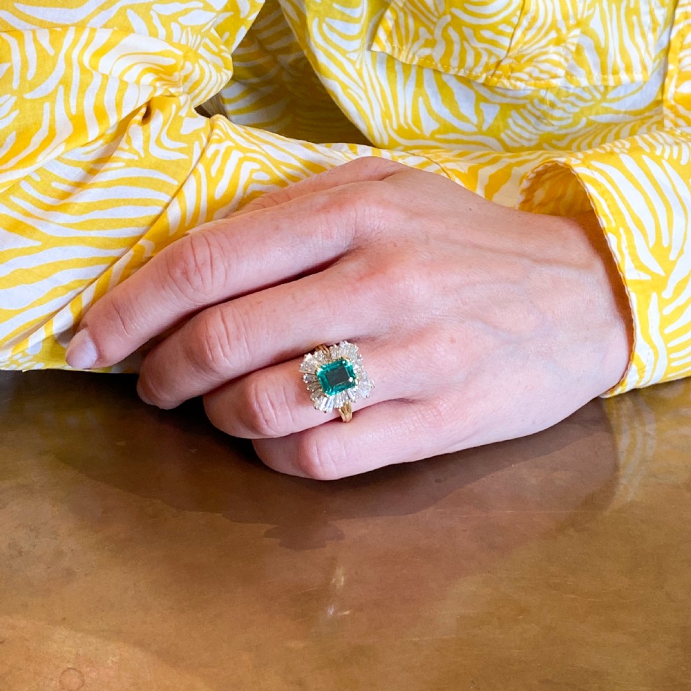 A yellow gold ballerina ring featuring a central rectangular emerald cut green emerald surrounded by a flared halo of baguette diamonds. View IX