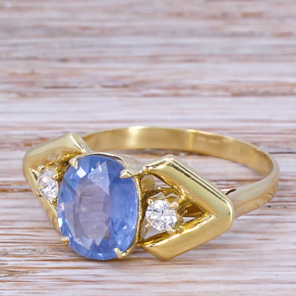A yellow gold ring featuring a central oval-cut light blue sapphire flanked by two round diamonds within geometric gold shoulders.