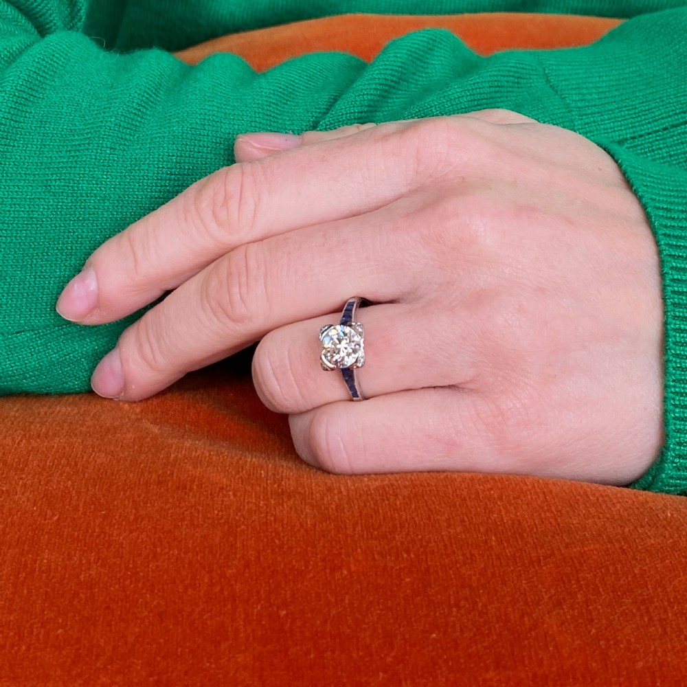 An Art Deco diamond solitaire ring set in white metal featuring calibre-cut sapphire shoulders and engraved metalwork. View VIII