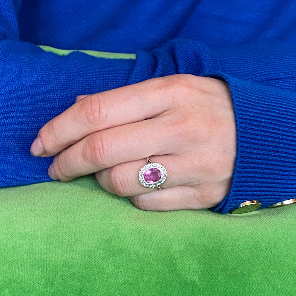 A mid-century ring featuring a central cushion-cut pink sapphire framed by a halo of small diamonds set in a white metal band with split shoulders. View VIII