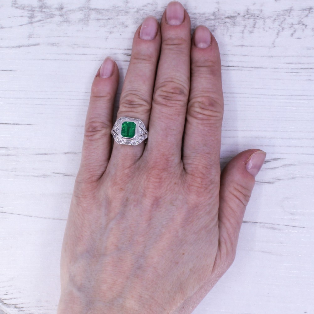 An Art Deco platinum ring featuring a central rectangular green emerald surrounded by a geometric halo of white diamonds with milgrain detailing. View VII