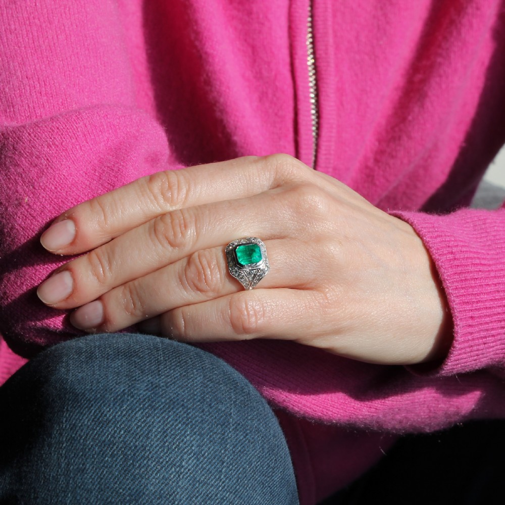 An Art Deco platinum ring featuring a central rectangular green emerald surrounded by a geometric halo of white diamonds with milgrain detailing. View X