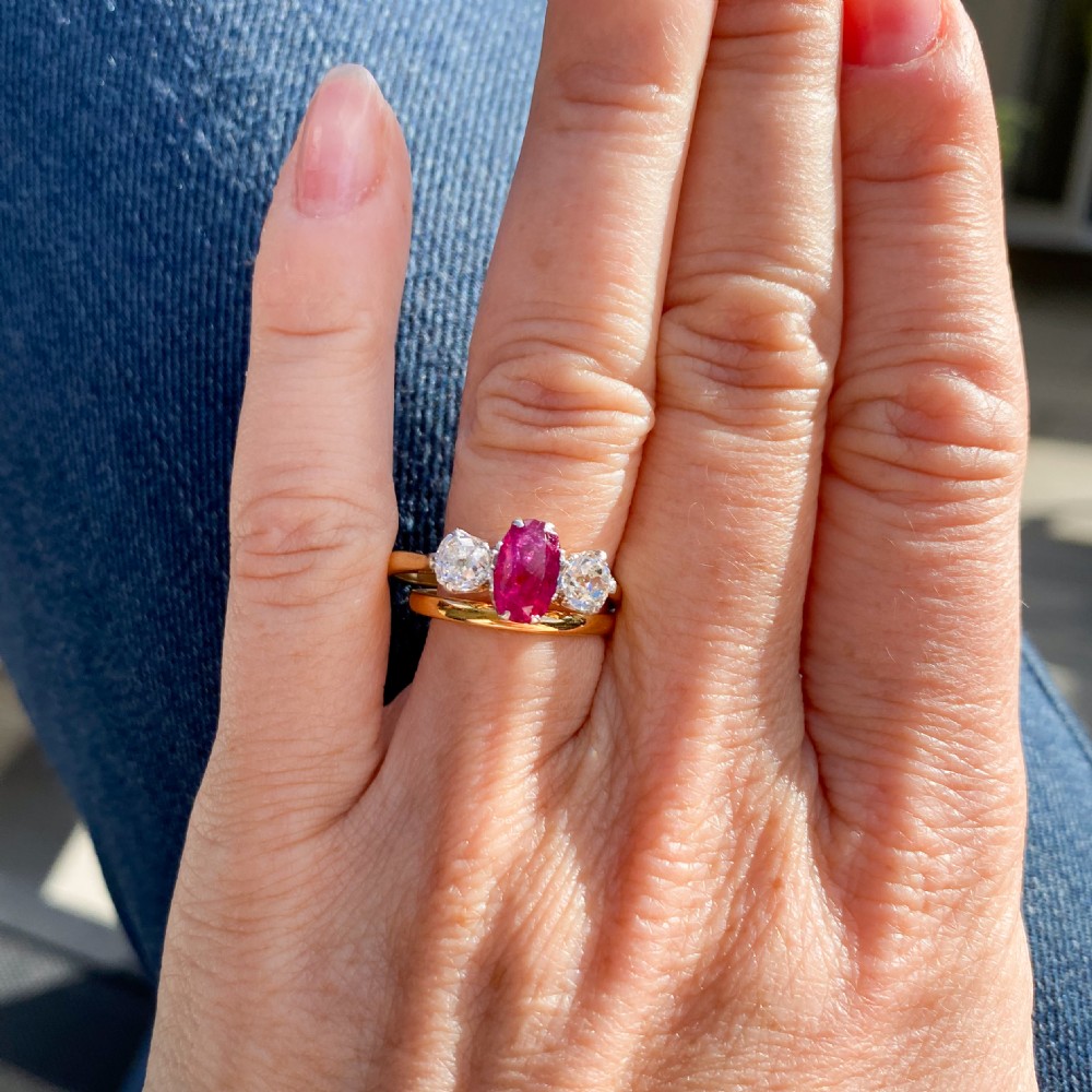 An Art Deco three-stone ring featuring a central oval red ruby flanked by two round diamonds on a yellow gold band with platinum claw settings. View IX