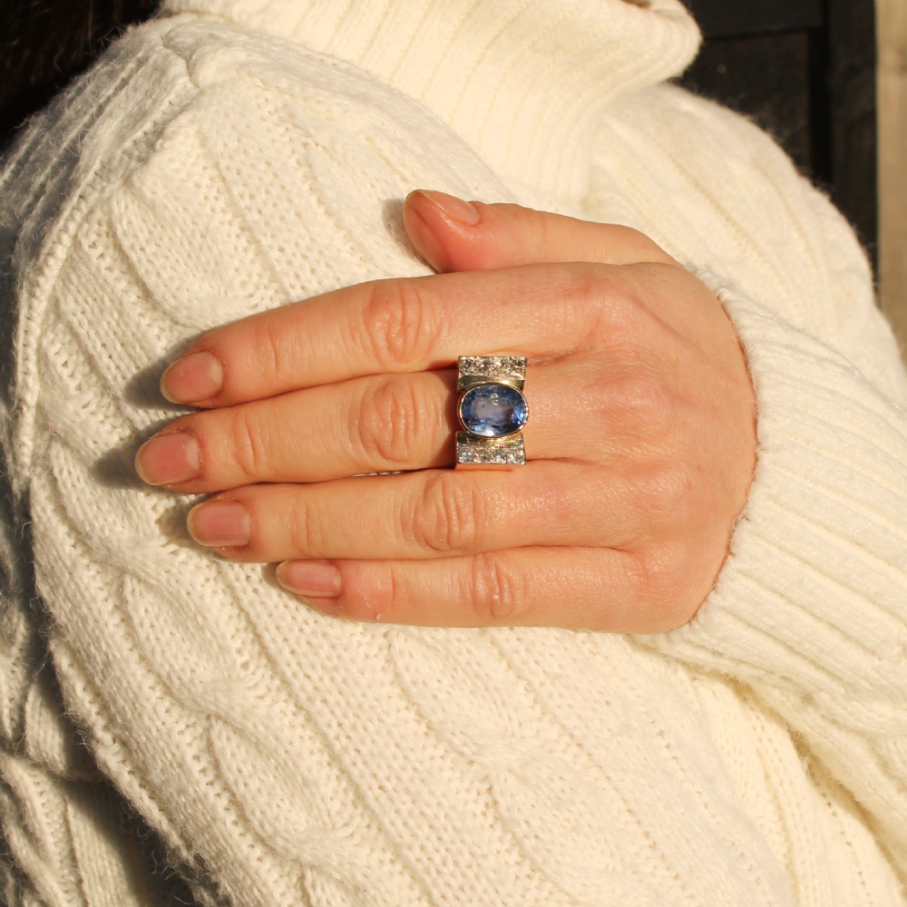 A mid-century gold ring featuring a central oval blue sapphire set in a wide band with diamond-set vertical shoulders. View VIII