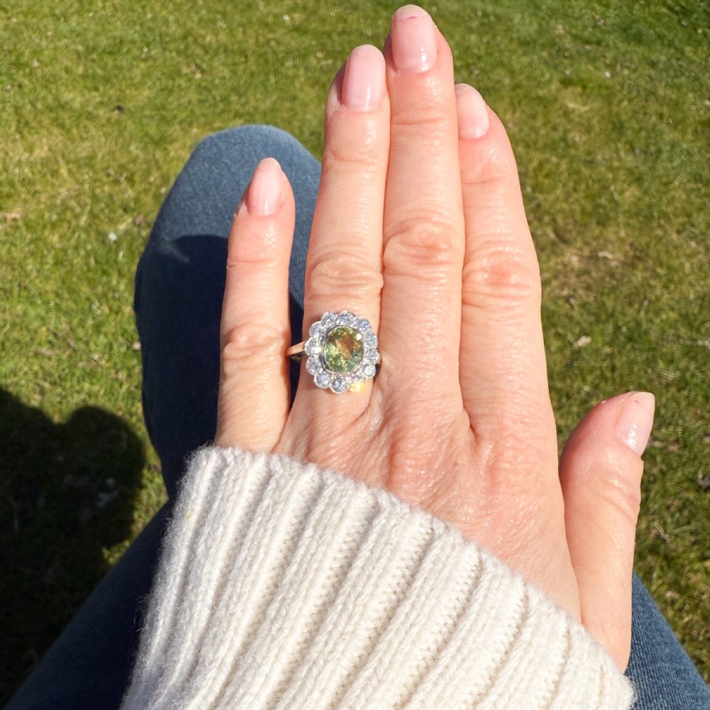 An Art Deco ring featuring an oval yellow-green chrysoberyl gemstone encircled by a halo of round diamonds set in platinum. View VI