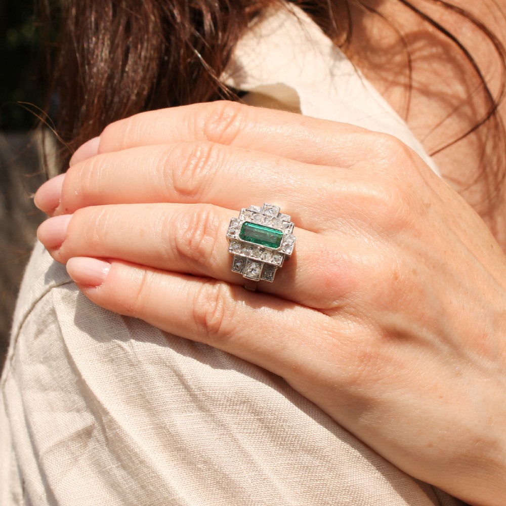 An Art Deco ring featuring a central rectangular emerald set within a stepped geometric frame of accent diamonds. View VI